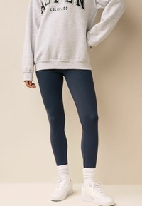 Felpa grigia con stampa "ASPEN COLORADO", abbinata a leggings blu scuro. Il modello indossa sneakers bianche e calzini bianchi a coste. Design semplice.