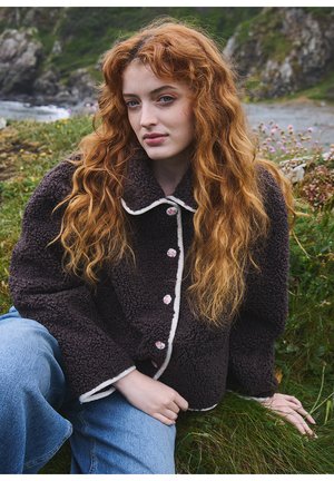 Jeune femme aux longs cheveux roux bouclés portant une veste polaire foncée et un jean bleu, assise sur l'herbe près d'une côte rocheuse.