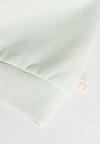 Poignet de sweat-shirt vert clair avec texture côtelée et une étiquette crème portant un graphique arc-en-ciel et le texte "Je suis heureux." Détail de tissu doux.