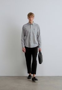 Svītrains garroku krekls ar pogājamu apkakli, valkāts kopā ar kroplētiem melniem biksēm un melniem apaviem. Tur melnu somu. Neitrāla fona.