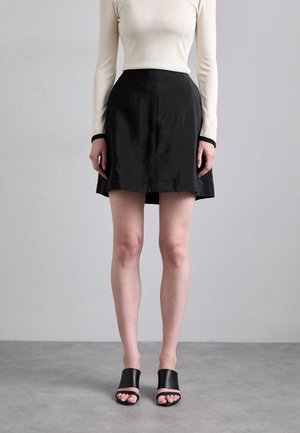 Femme portant un haut crème à manches longues, une jupe noire taille haute et des sandales noires à talons ouverts, debout sur un sol gris.