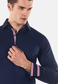 Camicia a maniche lunghe blu navy con accenti a carrè in plaid rosso e bianco sul colletto e sui polsini. Chiusura con bottoni, texture liscia.