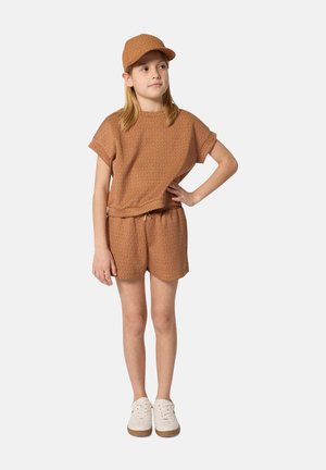 Young girl wearing brown cap, matching short-sleeve top and shorts, standing with one hand on hip and white sneakers on plain background.