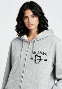 Felpa grigia con zip e cappuccio foderato in sherpa, con testo ricamato nero "Ai Amor" e un design a forma di cuore. Tasche frontali incluse.
