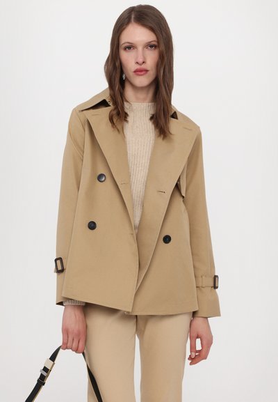 Veste beige à double boutonnage avec un large col, boutons noirs et poignets ajustables. Portée avec un pull beige clair et un pantalon.