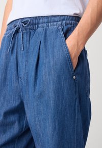 Denim jogger hlače u tamnoplavoj boji, s elastičnim pojasom i vezicom, bočnim džepovima te opuštenim krojem s nabranim detaljima.