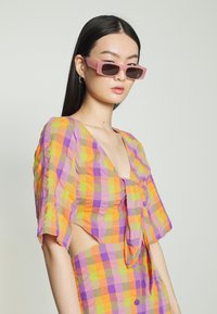 Conjunto colorido quadriculado em tons de laranja, roxo e verde, com mangas curtas e um design fluido, combinado com óculos de sol retangulares rosa.