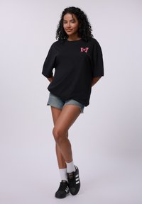 Svart oversized t-skjorte med et rosa sommerfuglgraphic, kombinert med denimshorts. Modellen har på seg svarte sneakers med hvite striper og hvite sokker.