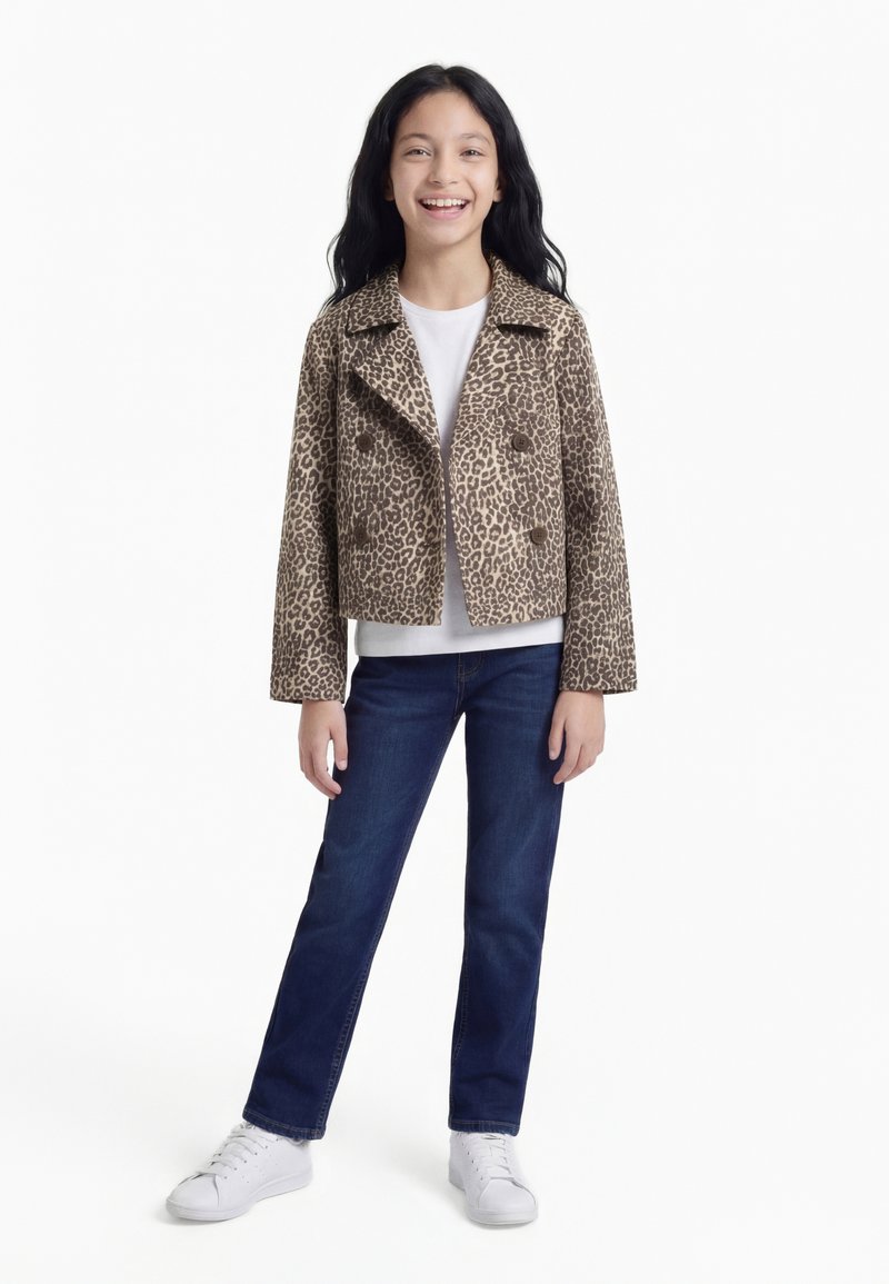 Šypsosi mergina, dėvinti leopardo rašto striukę, baltą marškinius, mėlynas džinsus ir baltus sportbačius, stovinti priešais paprastą baltą foną.