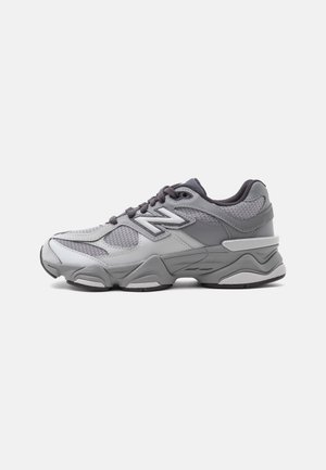 Baskets de sport grises et blanches avec panneaux en mesh et cuir, lacets gris foncé et semelle épaisse et rembourrée sur un fond clair uni.
