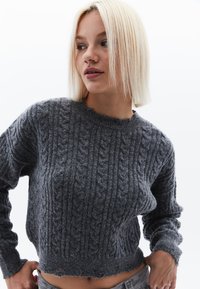 Grauer gestrickter Pullover mit Zopfmuster, rundem Halsauschnitt und gerippten Bündchen. Hat eine verkürzte Länge und einen leicht fransigen Saum.