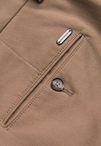 Tissu beige avec un tissage texturé, comportant une poche zippée et un bouton noir. Une étiquette en métal portant la mention "QUINTESSENCE" est attachée.