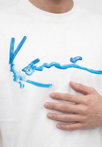 Mână sprijinită pe o cămașă albă cu un design abstract albastru lucios, în relief, pe piept.