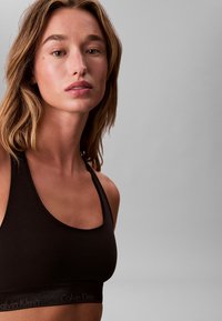 Schwarzer Sport-BH mit runder Ausschnittform und Racerback-Design. Mit einem Calvin Klein-Logo auf dem elastischen Bund. Glatte Textur.