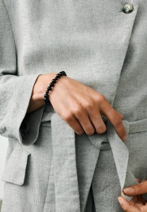 Person iført en lysegrå frakke, der spænder bæltet, med et sort perlearmbånd om håndleddet.