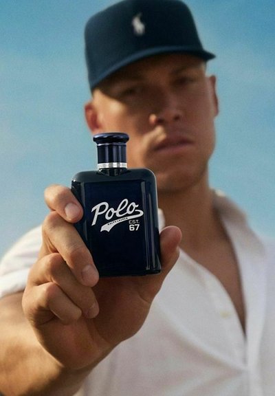 En man i en marinblå keps håller en mörkblå Polo-doftflaska framför en klar himmel, vilket främjar en fräsch doft.