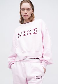 Sieviete, ģērbusies pasteļrozā Nike svārkā un biksēs ar bordo krāsas logo uzrakstu, stāv uz vienkāršas gaišas fona.