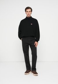 Μαύρος πλεκτός ζιβάγκο με άνετη εφαρμογή, συνδυασμένος με μαύρο παντελόνι και καφέ αθλητικά παπούτσια με λευκή σόλα. Μικρή λεπτομέρεια λογότυπου στο στήθος.