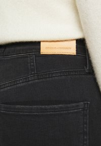 Los jeans de mezclilla negros tienen una etiqueta de cuero que dice "CITIZENS of HUMANITY". El primer plano muestra los detalles de la costura y los bolsillos.