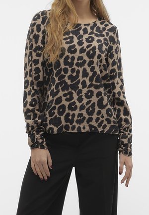 Femme portant un haut beige à manches longues avec un imprimé léopard noir et un pantalon noir à jambes larges, debout les mains détendues le long du corps.