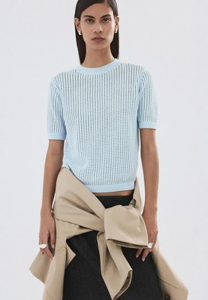 Kvinde iført lyseblå kortærmet striktop og beige jakke bundet om taljen, stående mod en ensfarvet lys baggrund.