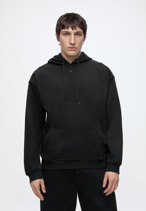 Giovane uomo con capelli corti e scuri, indossa una felpa nera semplice con coulisse e tasca frontale, in piedi davanti a uno sfondo bianco uniforme.