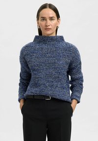 Maglione lavorato a maglia blu e bianco con colletto alto, maniche lunghe e una vestibilità rilassata, abbinato a pantaloni neri e una cintura nera.