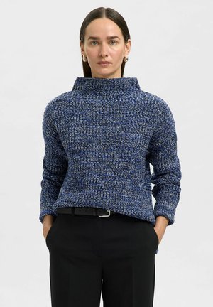 Maglione lavorato a maglia blu e bianco con colletto alto, maniche lunghe e una vestibilità rilassata, abbinato a pantaloni neri e una cintura nera.