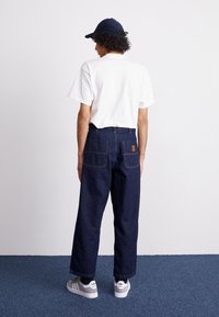 Donkerblauwe spijkerbroeken met een rechte pasvorm, zichtbare achterzakken en contrasterende stiksels. Gecombineerd met een wit T-shirt en grijze sneakers.