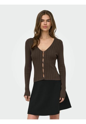 Femme portant un cardigan brun côtelé ajusté boutonné et une jupe noire trapèze, debout devant un fond clair uni.