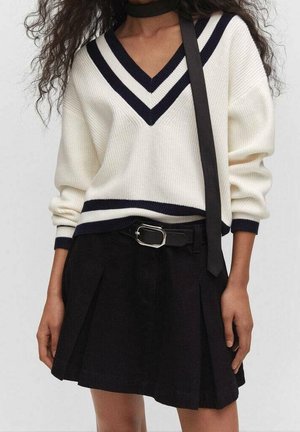 Kvinde iført en hvid V-hals sweater med sorte striber, en sort plisseret nederdel med bælte og et sort tørklæde, der hænger løst.