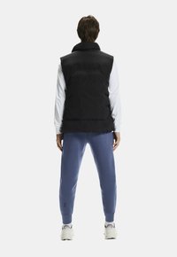 Czarna kamizelka puchowa z wysokim kołnierzem, zestawiona z niebieskimi spodniami dresowymi. Kamizelka ma poziome przeszycia, które dodają tekstury. Obuwie jest białe.