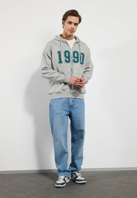 Grå hettejakke med teal "1990" trykk, brukt over en hvit t-skjorte, kombinert med lyseblå jeans og sneakers med mørkegrønne detaljer.
