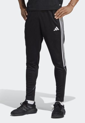 Homme portant un pantalon de survêtement Adidas noir avec des bandes blanches et un logo, des baskets noires et des chaussettes blanches, debout les mains sur les hanches.