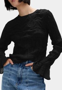 Schwarzes, strukturiertes Oberteil mit Rundhalsausschnitt und Glockenärmel. Getragen mit hellblauen Jeans. Der Stoff hat ein zerknittertes Muster.