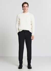 Weißer, strukturierter Strickpullover, kombiniert mit schwarzen, maßgeschneiderten Hosen und schwarzen Leder-Stiefeletten. Das Model steht vor einem neutralen Hintergrund.