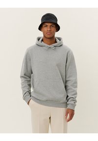 Gray cotton hoodie with a drawstring and front pocket, paired with beige pants and a black bucket hat, set against a light background.