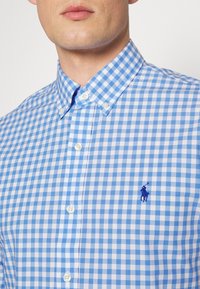 Blå och vit rutmönstrad skjorta med knäppkrage, som har vita knappar och en liten blå broderad logotyp på bröstet.