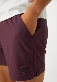 Ljubičaste kratke hlače od glatkog materijala, s bočnom džepom i crnom logotipom na rubu. Dizajn je jednostavan i prilagođen tijelu.