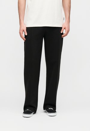 Homme portant un pantalon noir ample et des baskets en toile noires avec des semelles blanches, debout devant un fond gris clair.