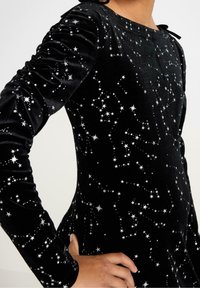 Sort velourkjole med lange ærmer, der har sølvstjerne mønstre. Designet inkluderer draperet skulderdetalje og en rund halsudskæring.