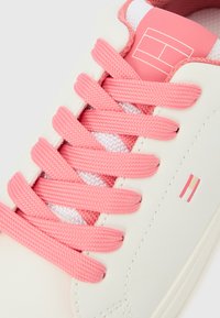 Zapatilla blanca con cordones rosas, lengüeta rosa con un logo geométrico blanco y una pequeña "H" rosa y beige en el lateral.