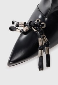 Zapato de cuero negro con punta afilada, que presenta detalles metálicos y un intrincado sistema de cordones con herrajes de estilo muelle y correas de cuero.
