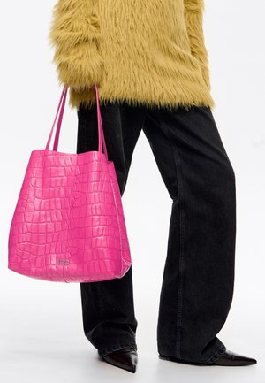 Osoba ubrana w czarne spodnie o szerokich nogawkach i żółty, puszysty sweter, trzymająca jaskraworóżową, teksturowaną torbę typu tote z długimi uchwytami.