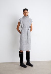 Grau gestricktes Kleid mit hohem Kragen, Taillengürtel, kurzen Ärmeln und seitlichen Schlitzen. Kombiniert mit schwarzen knielangen Stiefeln. Glatte Textur.