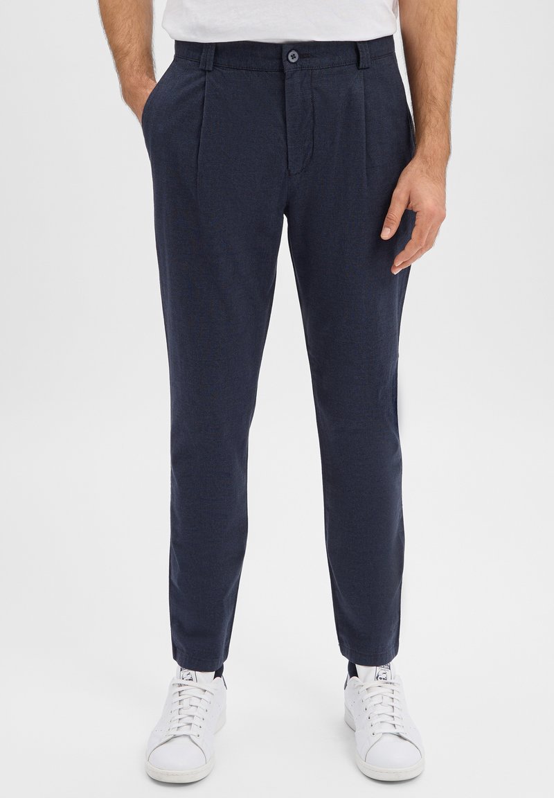 Granatowe spodnie o gładkiej fakturze, z przodu plisowane i zapinane na guzik. Połączone z białymi sneakersami dla casualowego wyglądu.