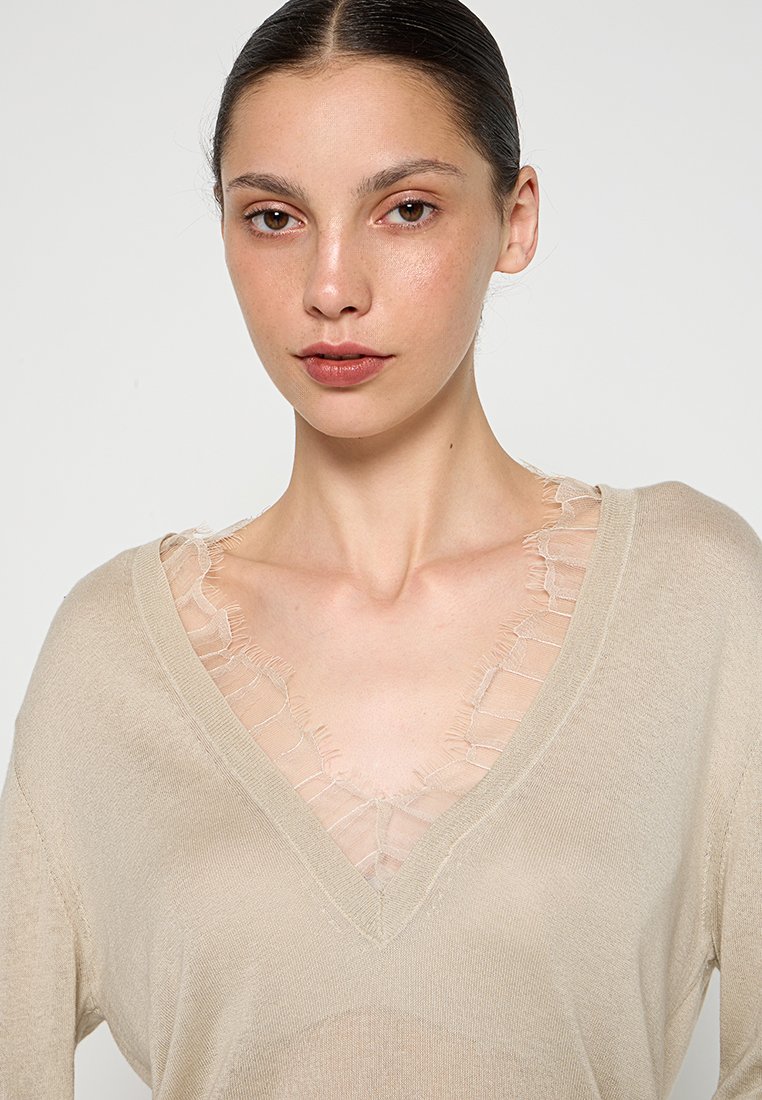 Jonge vrouw met donker haar in een beige V-hals trui, gedragen over een delicate doorzichtige top met ruches, neutrale achtergrond.