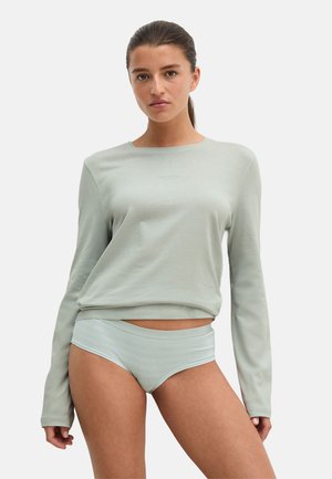 Femme aux cheveux foncés en queue de cheval portant un haut à manches longues vert clair et des sous-vêtements assortis verts clairs, debout devant un fond uni.