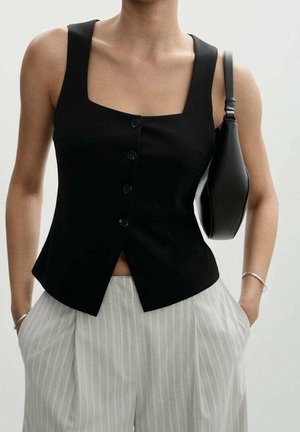Femme portant un haut noir sans manches boutonné, un pantalon gris rayé, et portant un sac à épaule noir avec les mains dans les poches.