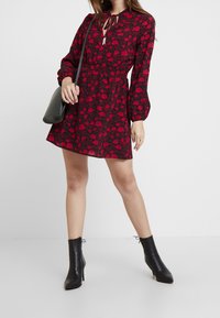 Robe à motifs floraux rouge et noir avec de longues manches bouffantes, taille élastique et un détail de nœud au niveau du décolleté. Portée avec des bottines noires.